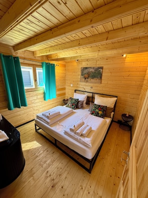 A wooden cabin bedroom with a bed, curtains, a painting, and a small table.