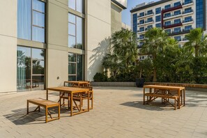 An outdoor patio area with wooden tables and chairs, surrounded by a building with large windows and a balcony. - HAVN - Prime Studio in Torino Arjan in Garden View (Dubai)