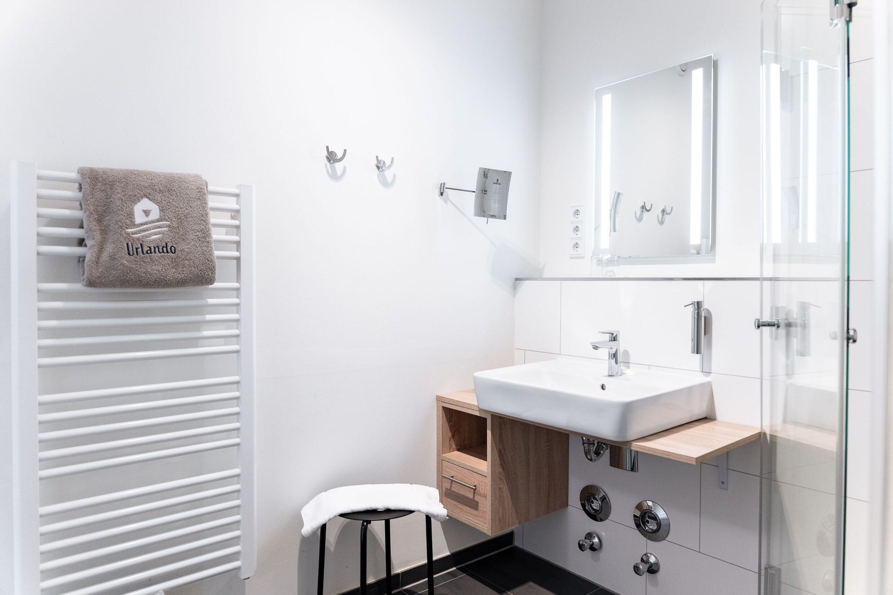 Un baño moderno con un lavamanos blanco, una toalla con un logotipo, un espejo y un box de ducha de vidrio.