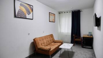 A room with a brown sofa, a small white table, a chair, and two framed artworks on the wall.