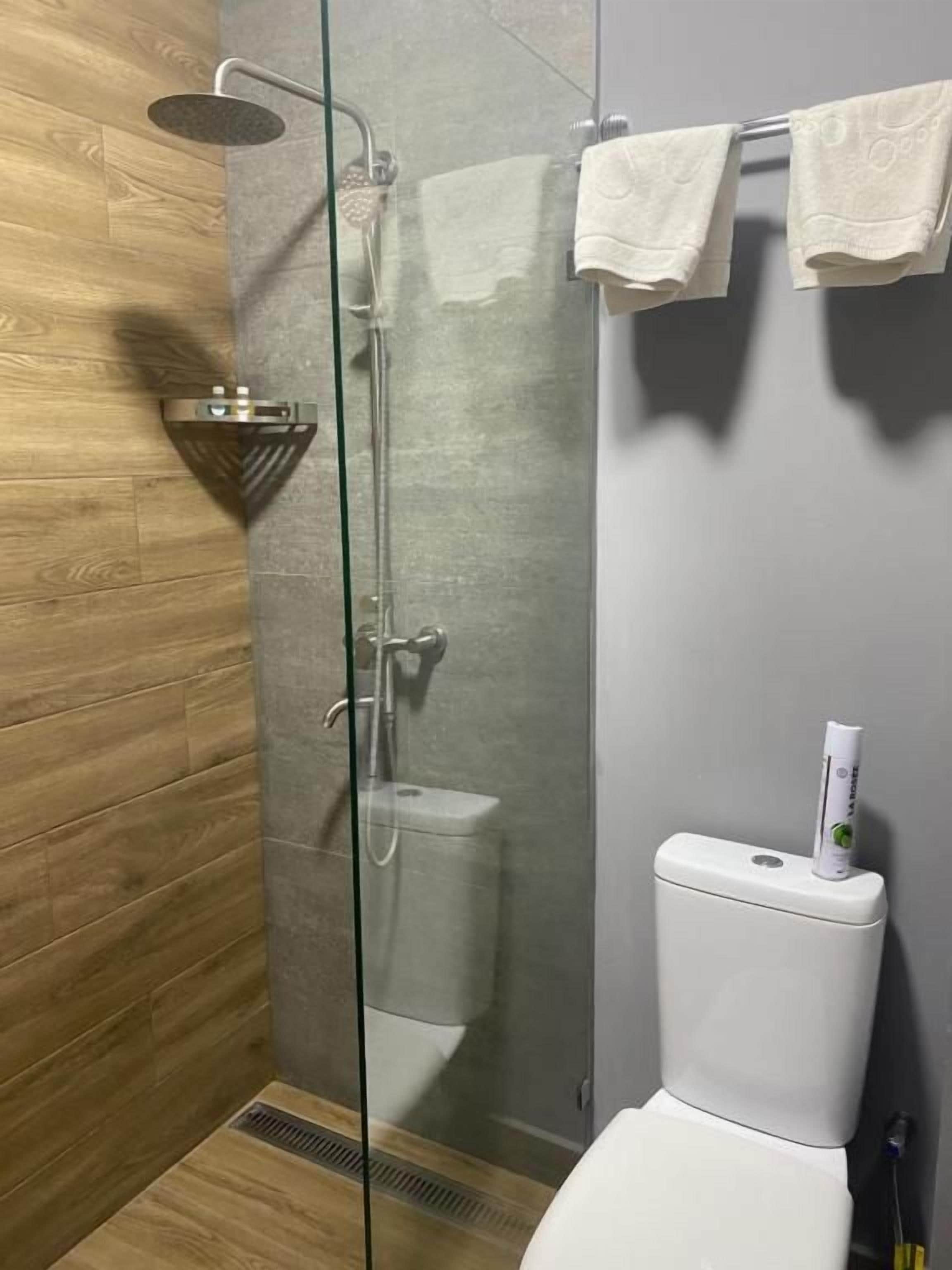 Et moderne badeværelse med en brusekabine i glas, et hvidt toilet og trægulv.