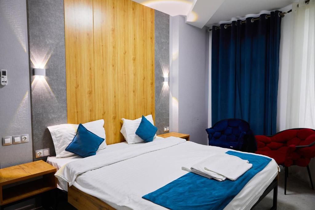 A hotel room with a wooden headboard, a bed with white and blue bedding, a blue throw blanket, a red chair, and a blue curtain.