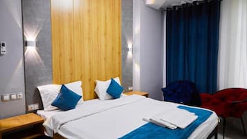 Habitación de hotel con cabecera de madera, cama con ropa de cama blanca y azul, una manta azul, una silla roja y una cortina azul.