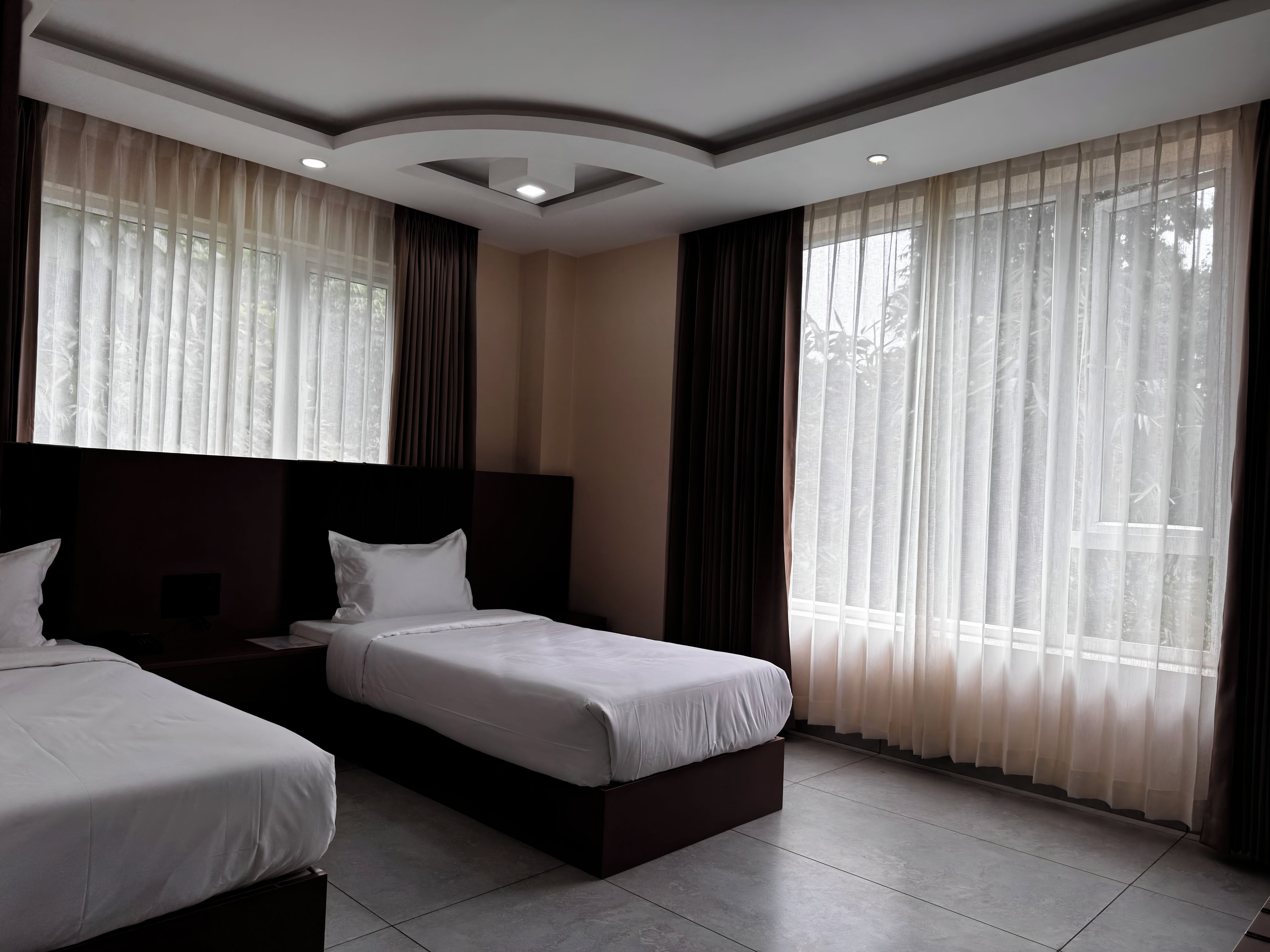 Una habitación de hotel con dos camas, un ventanal con cortinas translúcidas y un diseño moderno en el techo.