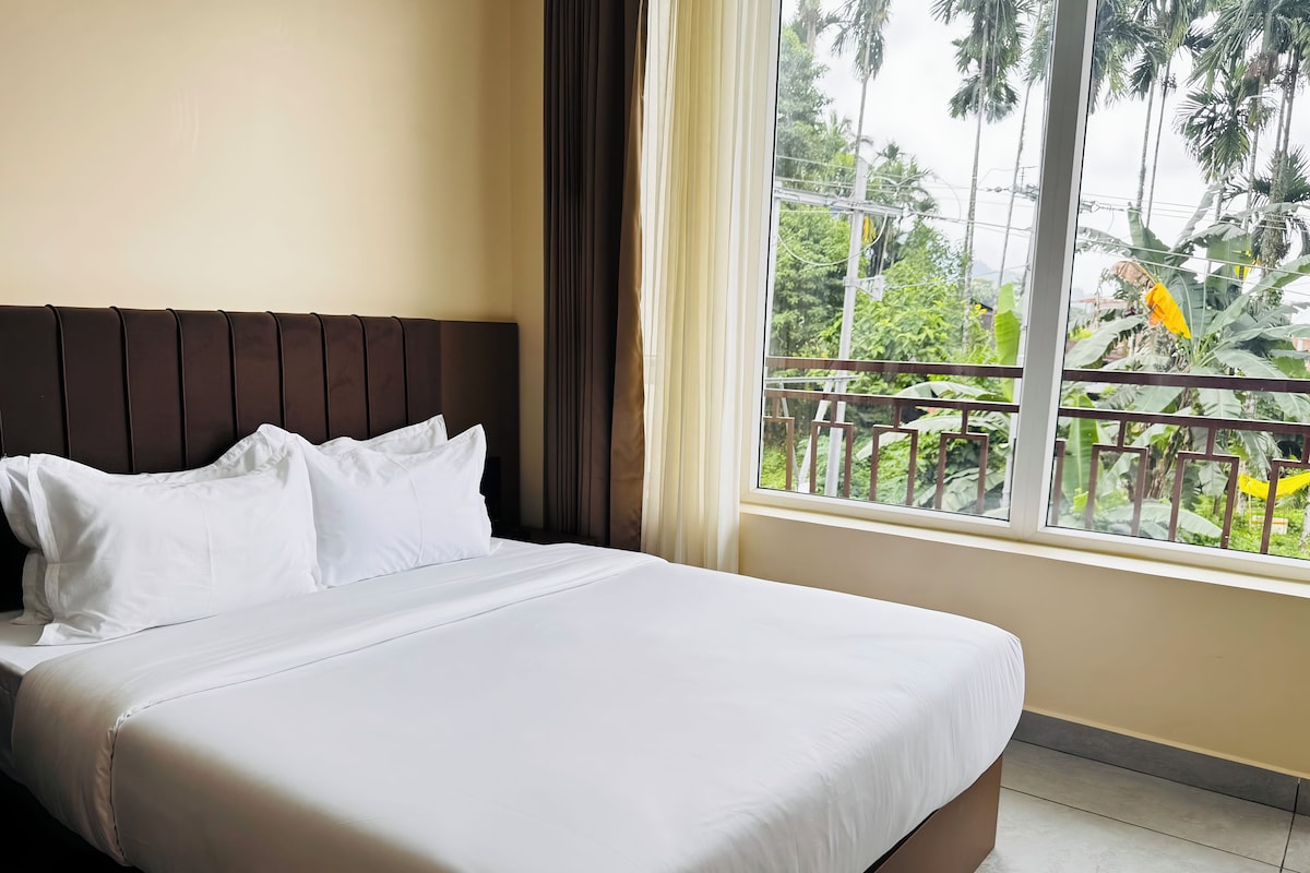 A hotel room with a bed, a window with a view of greenery, and a curtain.
