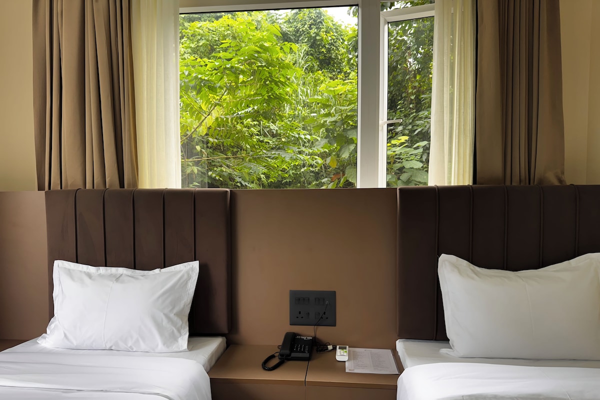 A hotel room with two beds, a bedside table with a phone and papers, and a window with a view of greenery.
