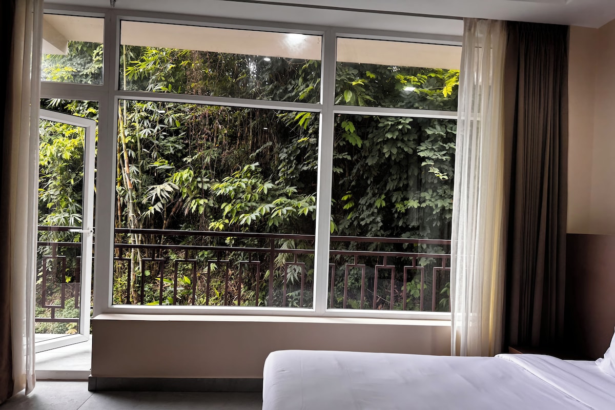 A hotel room with a large window, a bed, and a view of dense foliage.
