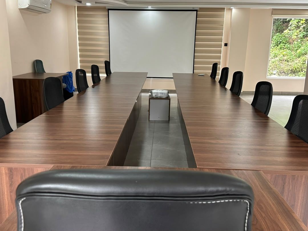 Un salón de conferencias con una larga mesa, sillas, una pantalla de proyector y vistas al exterior con zonas verdes.