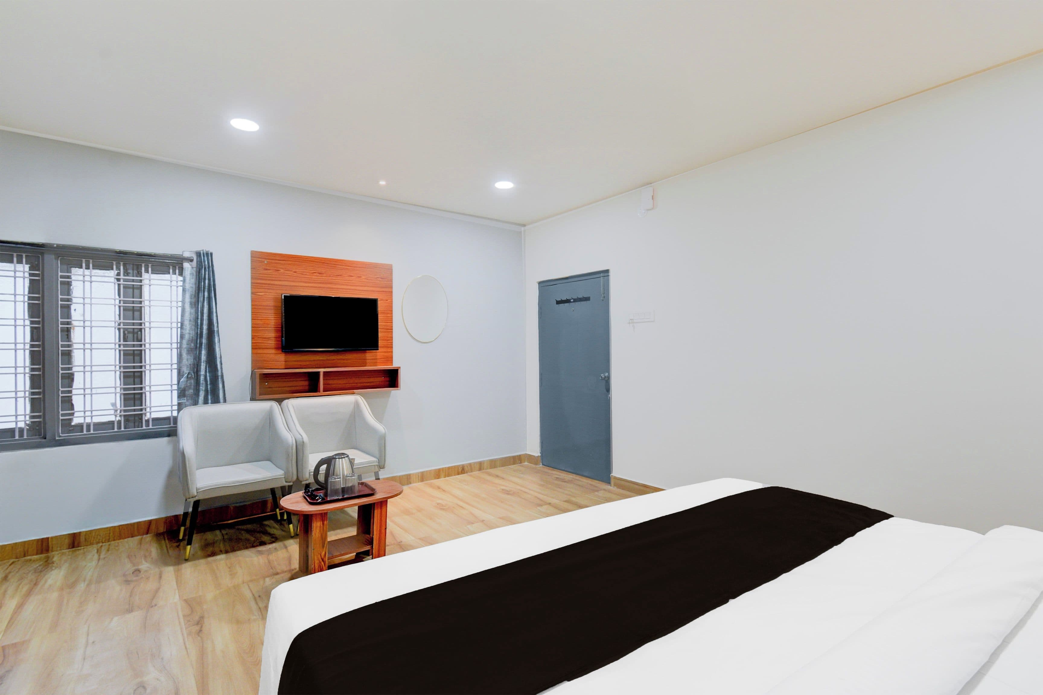 A hotel room with a bed, a TV mounted on the wall, a chair, a small table with a kettle, and a window with curtains.