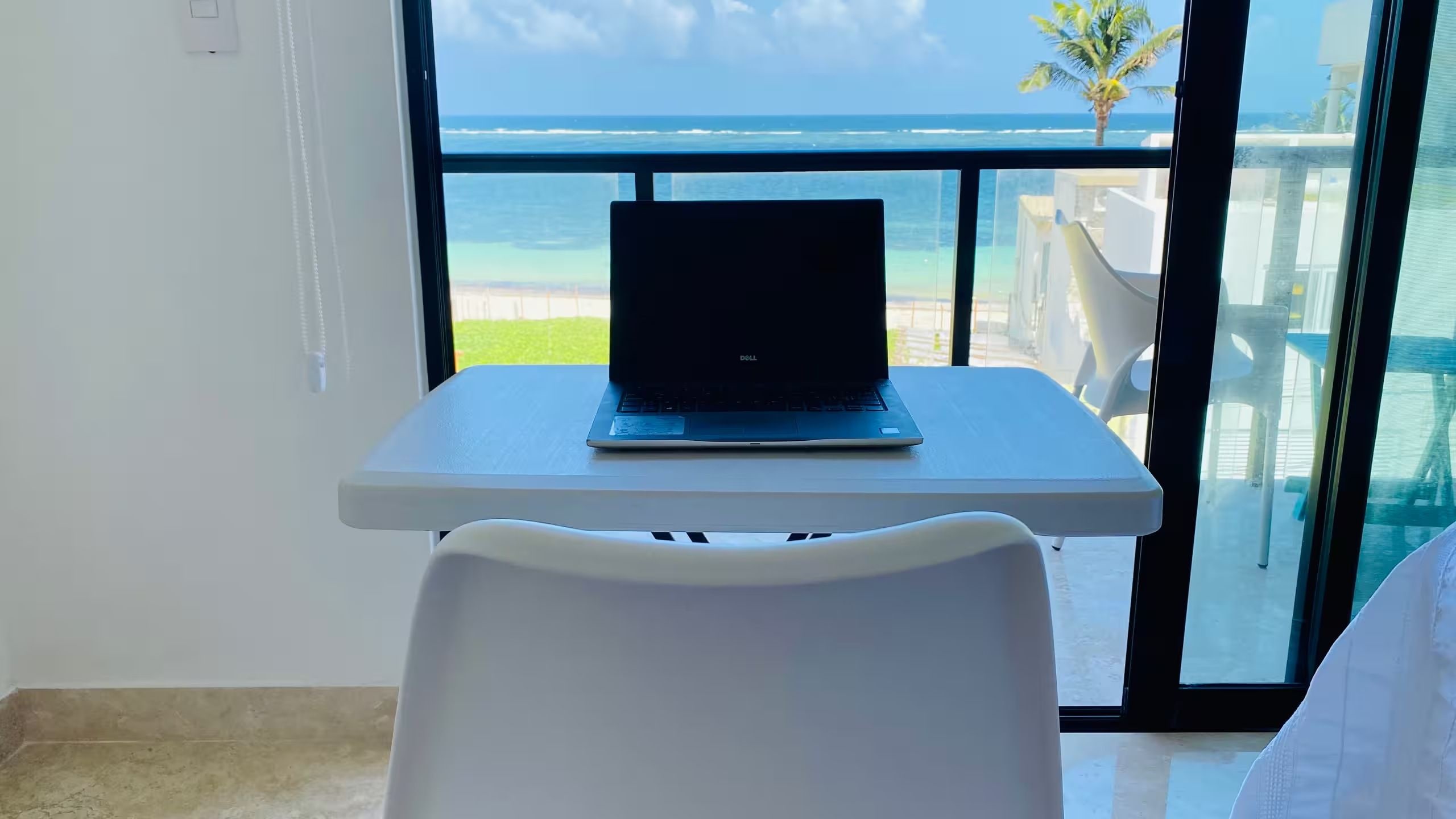 Un ordinateur portable est posé sur une petite table, avec vue sur l’océan et un palmier.