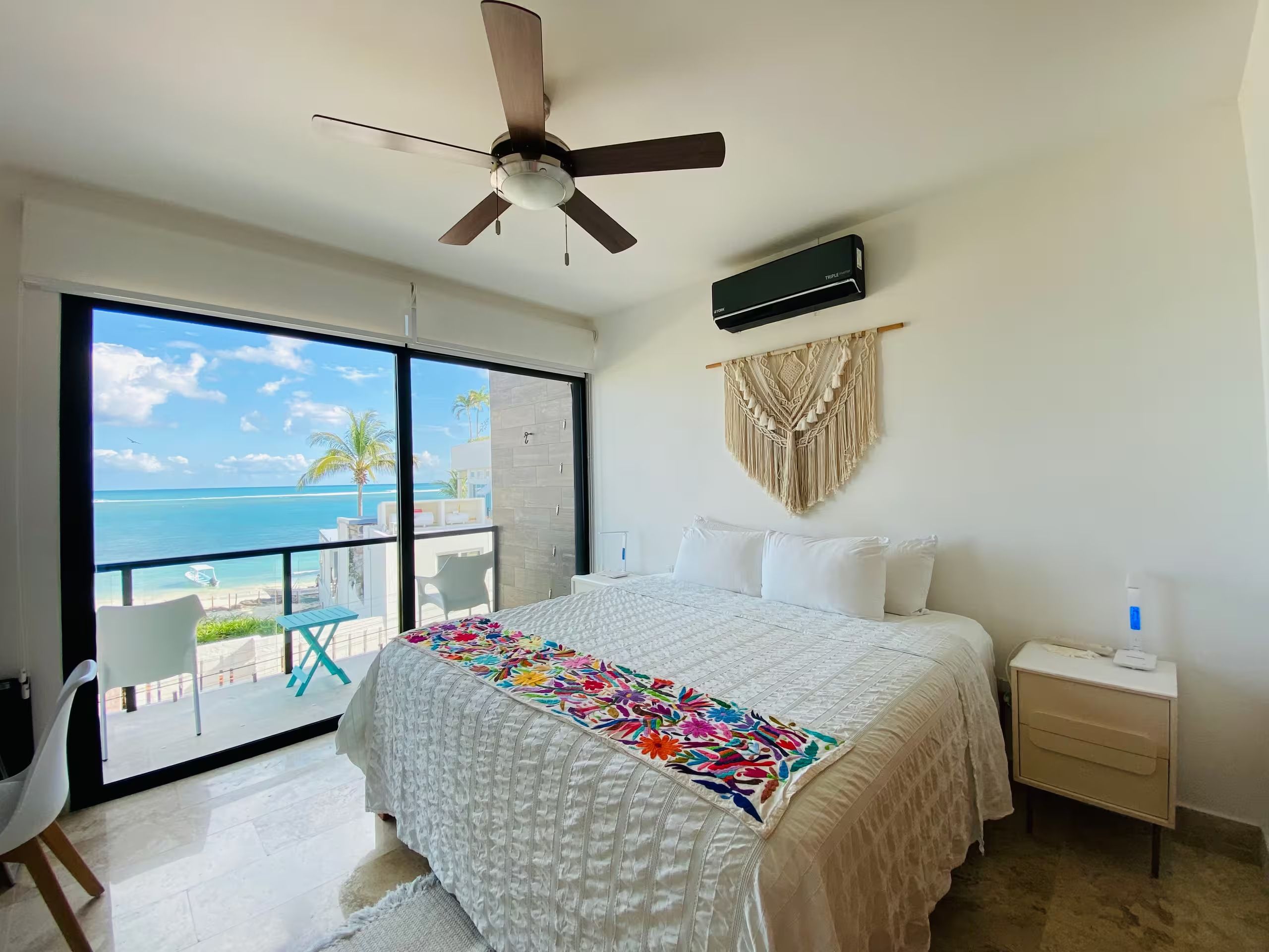 A hotel room with a bed, a ceiling fan, a balcony with a view of the ocean, and a macrame wall hanging.