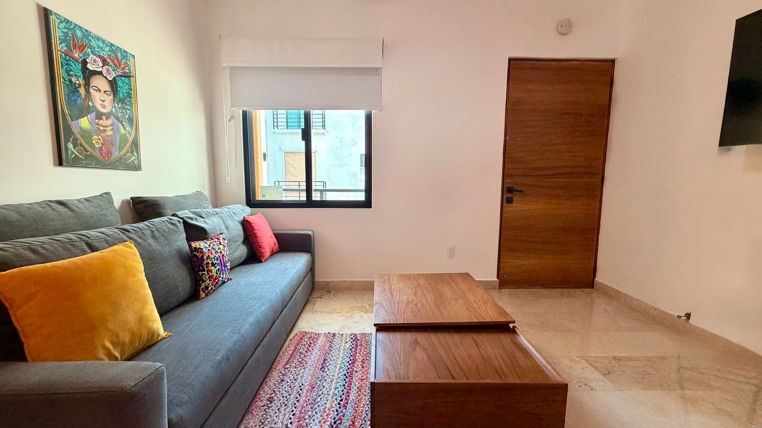 Un salon avec un canapé gris, des coussins colorés, une table basse en bois, un tapis à motifs et un tableau accroché au mur.