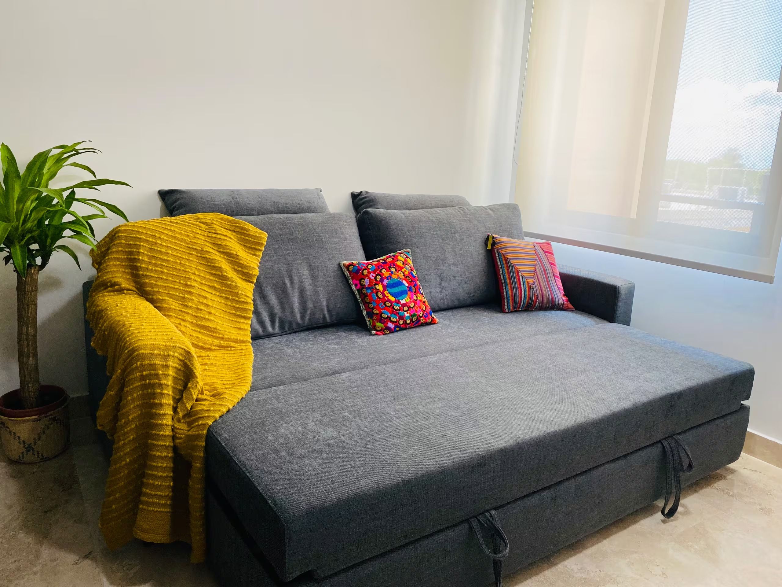 Um sofá cinzento com um cobertor amarelo, duas almofadas coloridas e um vaso com planta.