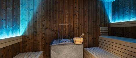A sauna with wooden walls and benches, a central stone platform, and a small wooden bucket.