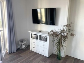 Ein Zimmer mit einem an der Wand befestigten Flachbildfernseher, einer weißen Kommode mit Schubladen, einem Ventilator und einer Vase mit Trockenpflanzen.