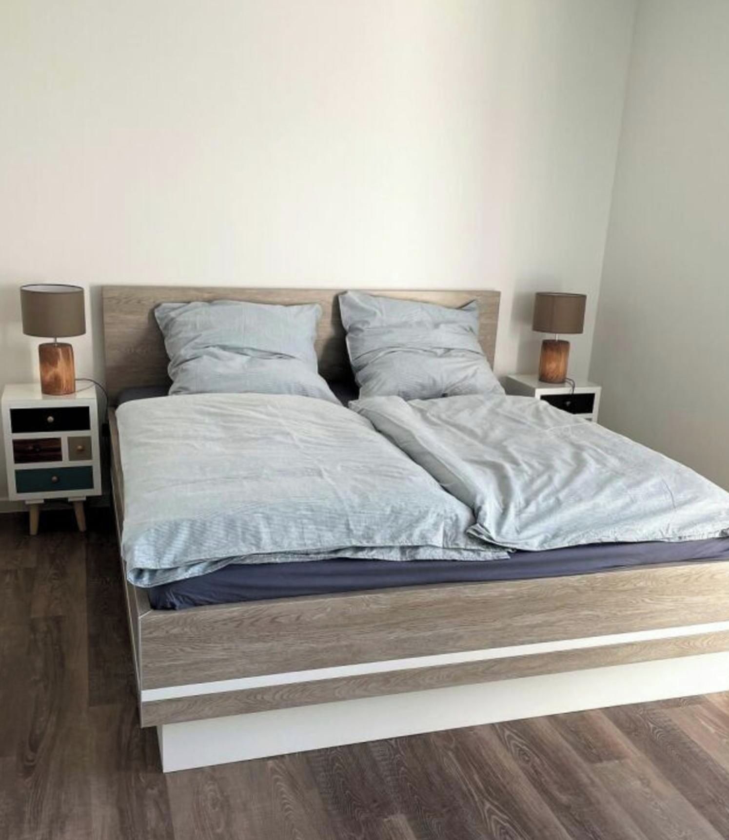 Un letto matrimoniale con struttura in legno e biancheria grigia, affiancato da due comodini con lampade, in una stanza con pavimento in legno.