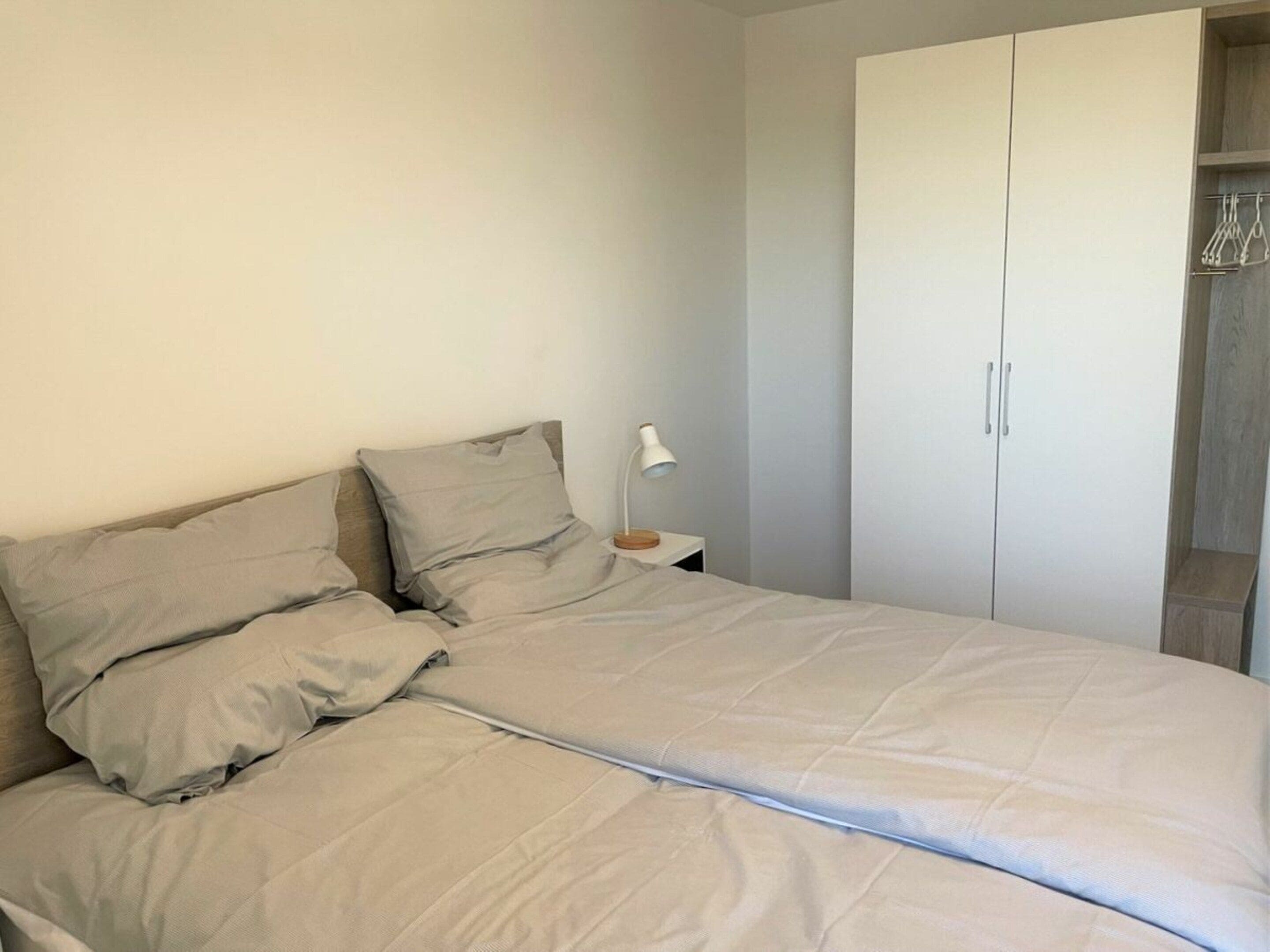 En seng med hvid dyne og puder, et natbord med lampe, og et garderobeskab med bøjler.