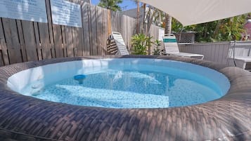 A hot tub surrounded by a wooden fence, with lounge chairs and an umbrella in the background.