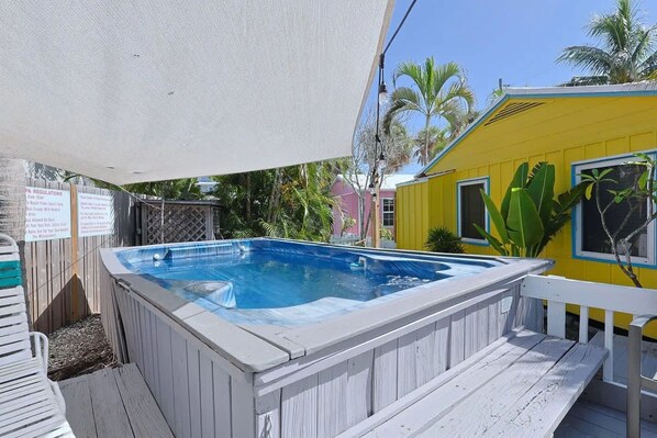 A hot tub adjacent to a wooden deck with a white railing, surrounded by lush greenery and a yellow building.