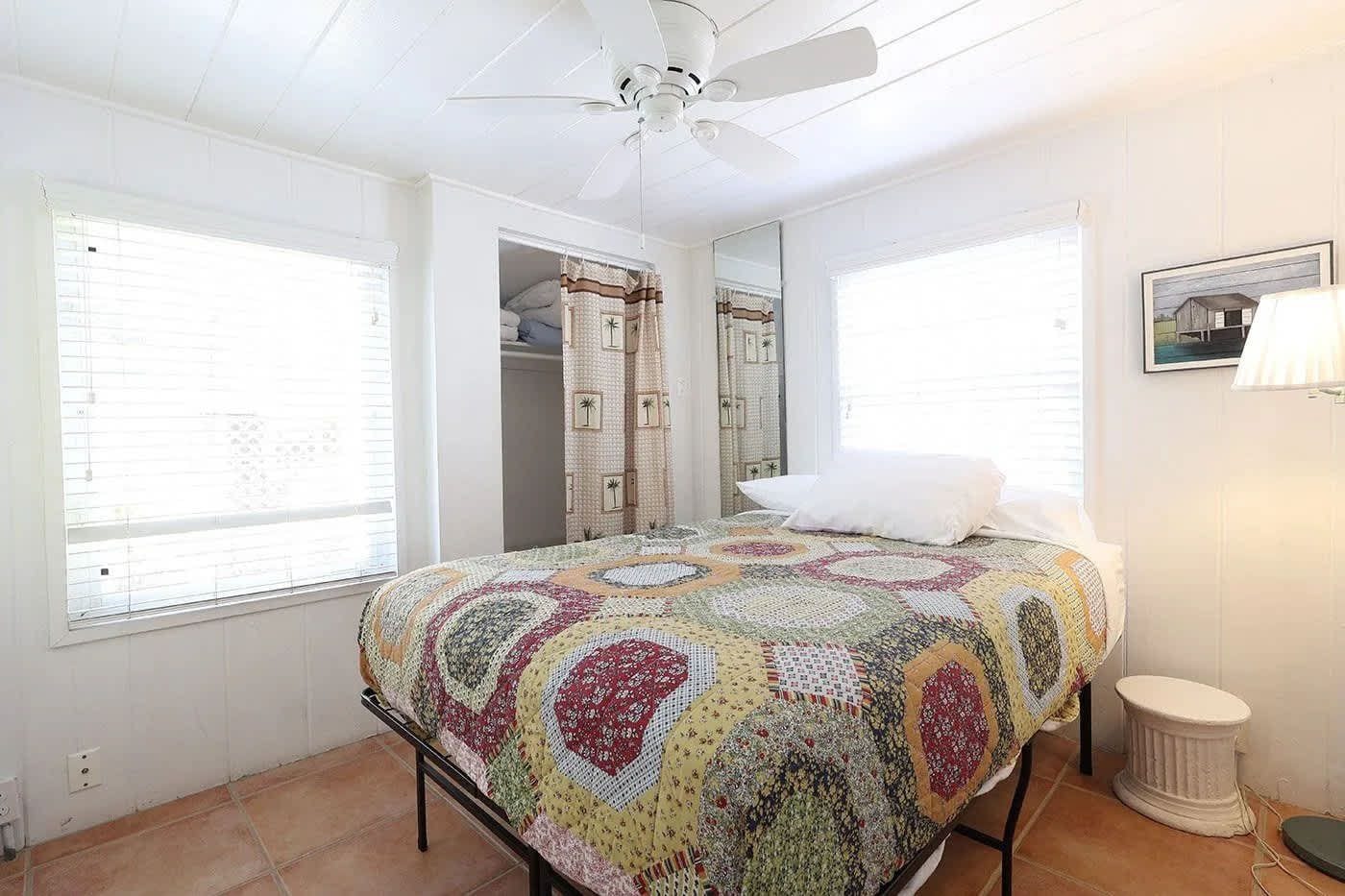 Un dormitorio con cama, ventilador de techo, ventana, espejo y un taburete pequeño.