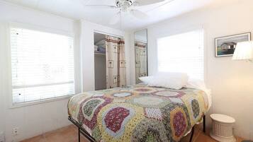 Un dormitorio con cama, ventilador de techo, ventana, espejo y un taburete pequeño.