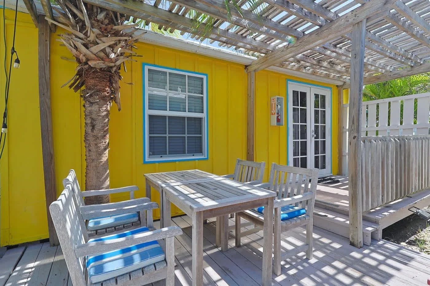 A covered outdoor area with a wooden table and chairs, a white door, and a yellow wall.