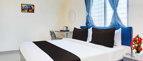 A hotel room with a bed, a desk, a chair, a nightstand, a vase with flowers, and a painting on the wall.