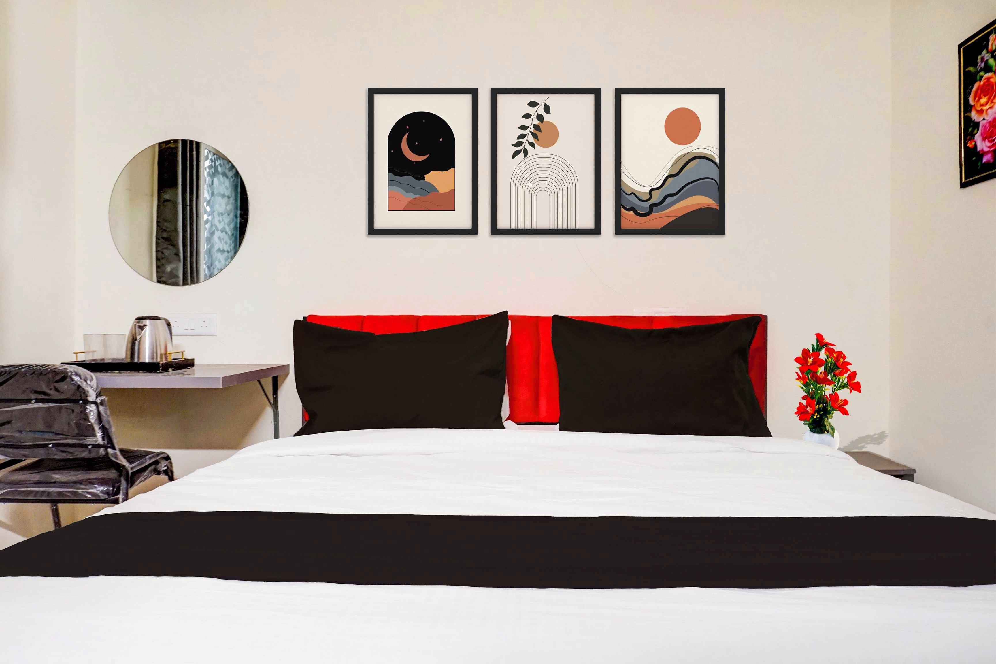 A hotel room with a bed, a desk, a chair, a round mirror, and three framed artworks on the wall.