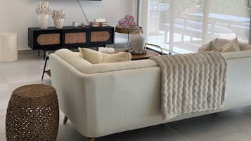 Un salon moderne doté d’un grand téléviseur à écran plat, d’un canapé blanc, d’une petite table d’appoint en osier et d’un luminaire suspendu au plafond.