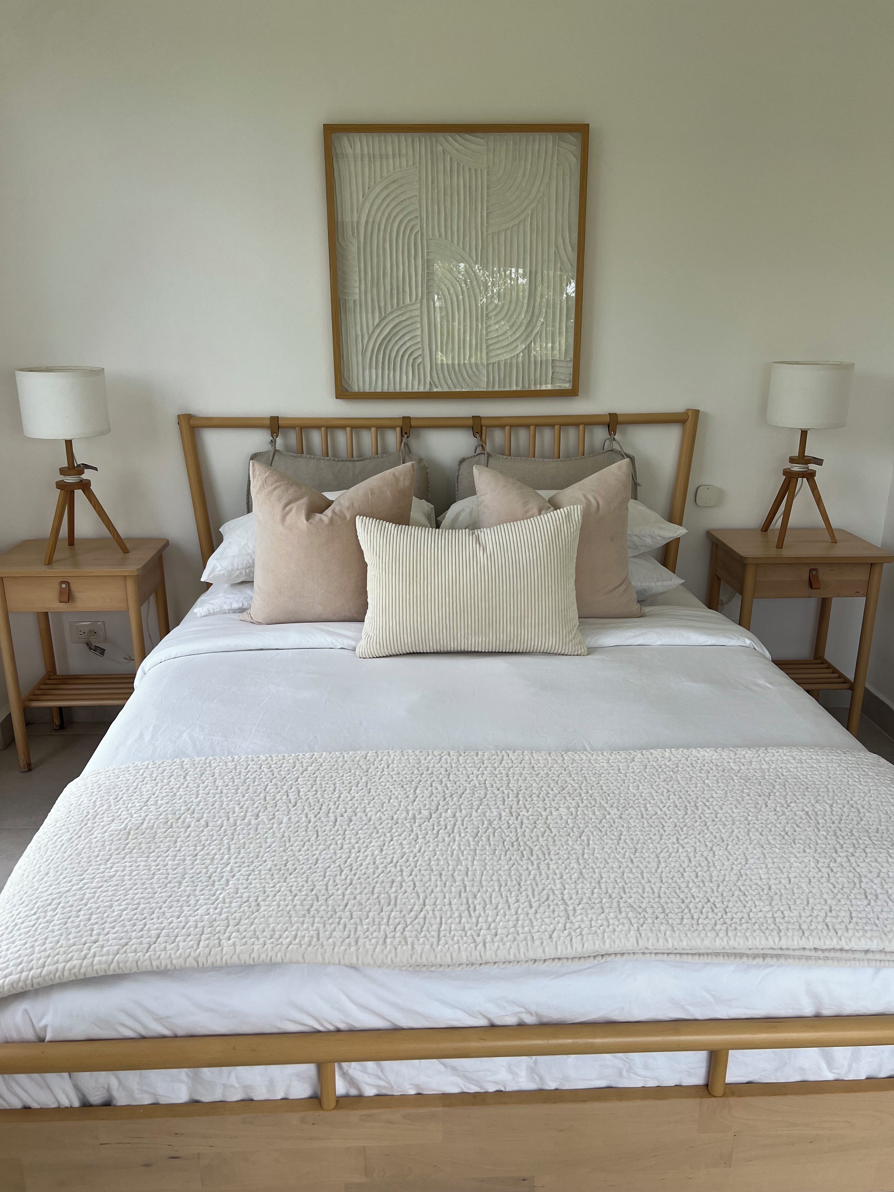 A neatly made bed with a white duvet and several pillows, flanked by two bedside tables with lamps, in a room with a wooden headboard and a framed artwork on the wall.