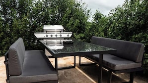 Une terrasse extérieure avec une table à plateau en verre, deux bancs rembourrés et un barbecue en acier inoxydable.