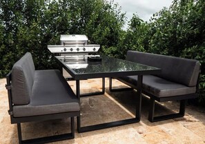 Un patio al aire libre con una mesa de vidrio, dos bancos acolchados y una parrilla de acero inoxidable.
