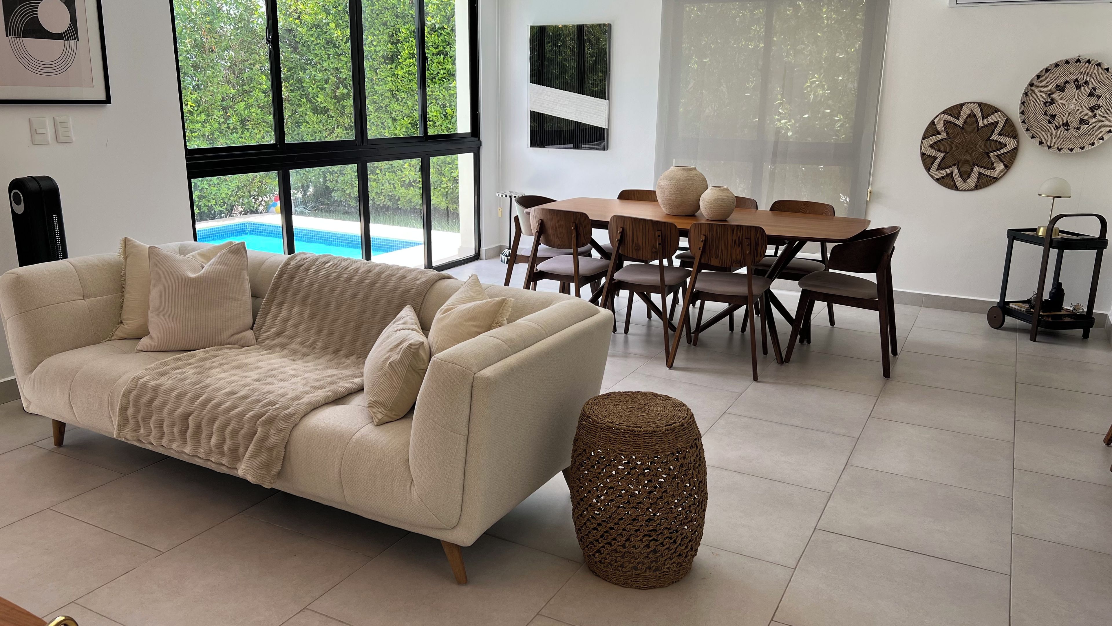 A modern living room with a beige sofa, a dining table with chairs, and a large window with a view of greenery.