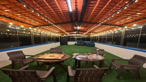 Un espacio interior con techo de listones de madera, rodeado de muros de cristal. Hay mesas con sillas y un banco.