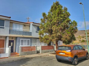Een gebouw met twee verdiepingen, een balkon, een geparkeerde oranje auto en een grote dennenboom.