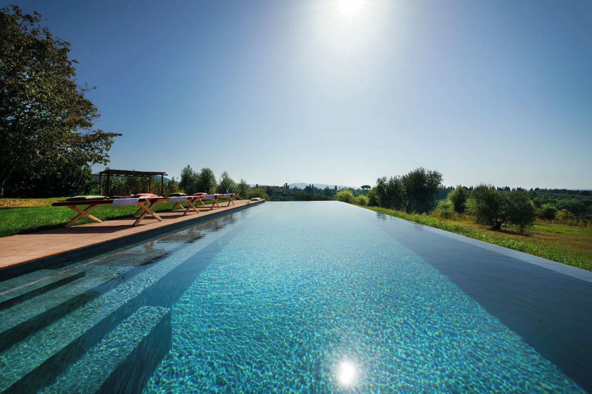 A clear swimming pool with steps leading into it, surrounded by greenery and a clear sky.