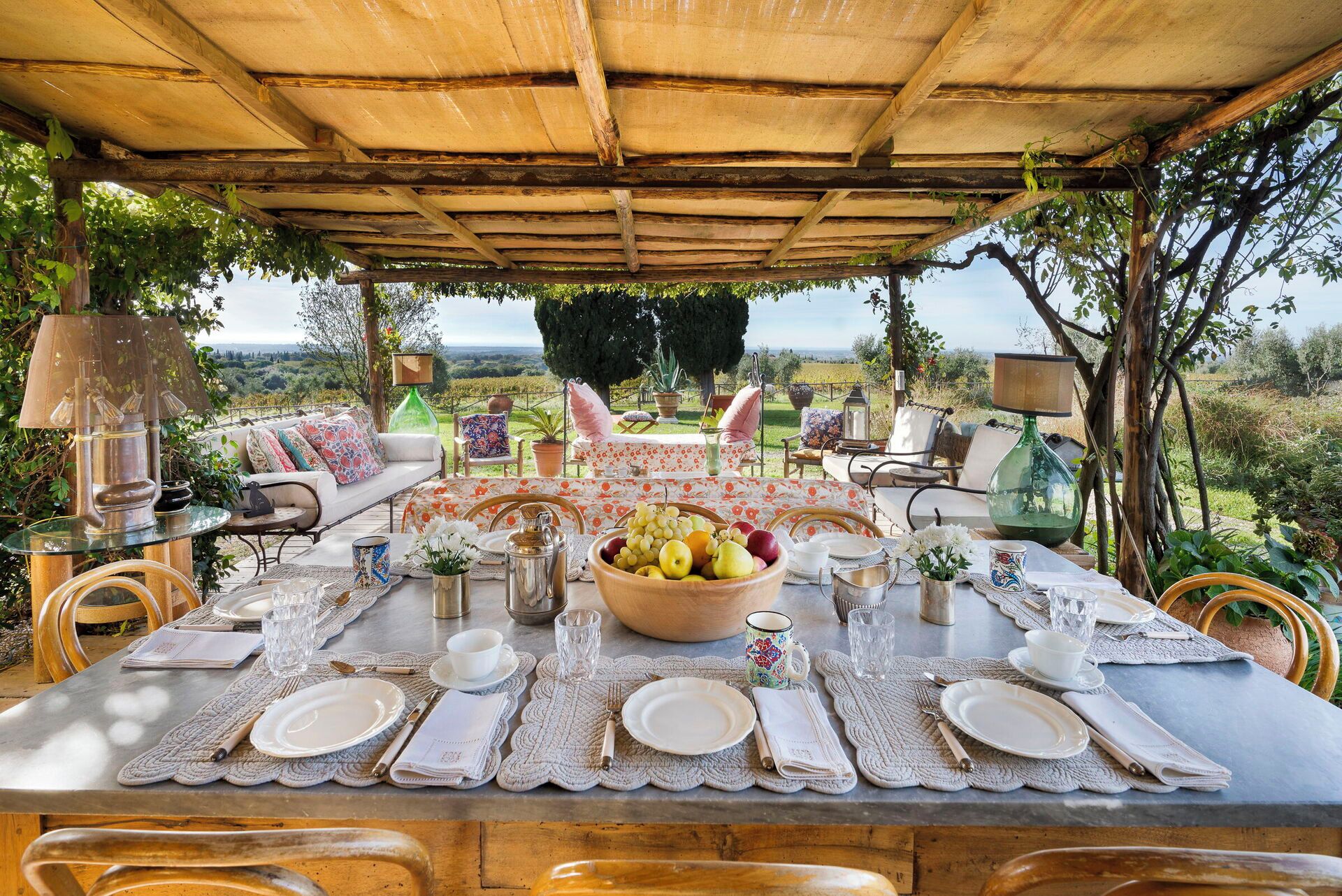 An outdoor dining area with a table set for four, surrounded by chairs and a sofa, overlooking a scenic landscape.