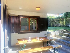 Ein Essbereich im Freien mit Tisch und Bänken, ein Fenster mit Fensterläden sowie eine Steintreppe, die in einen Garten führt.