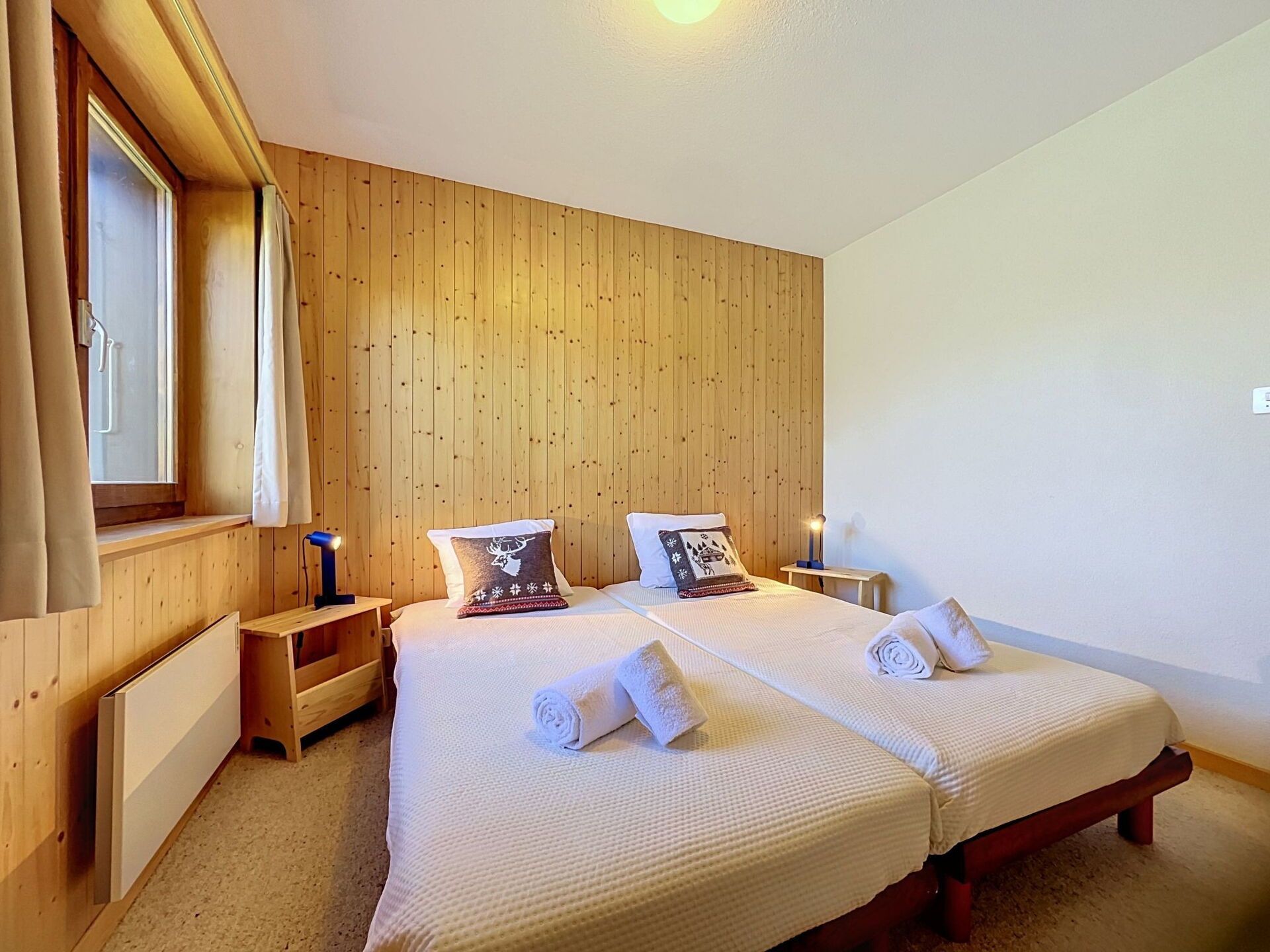 A double room with two beds, wooden panel walls, a window with curtains, bedside lamps, and a radiator.
