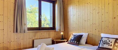 A room with wooden panel walls, a double bed with white bedding, two pillows, a nightstand with a lamp, and a window with curtains.
