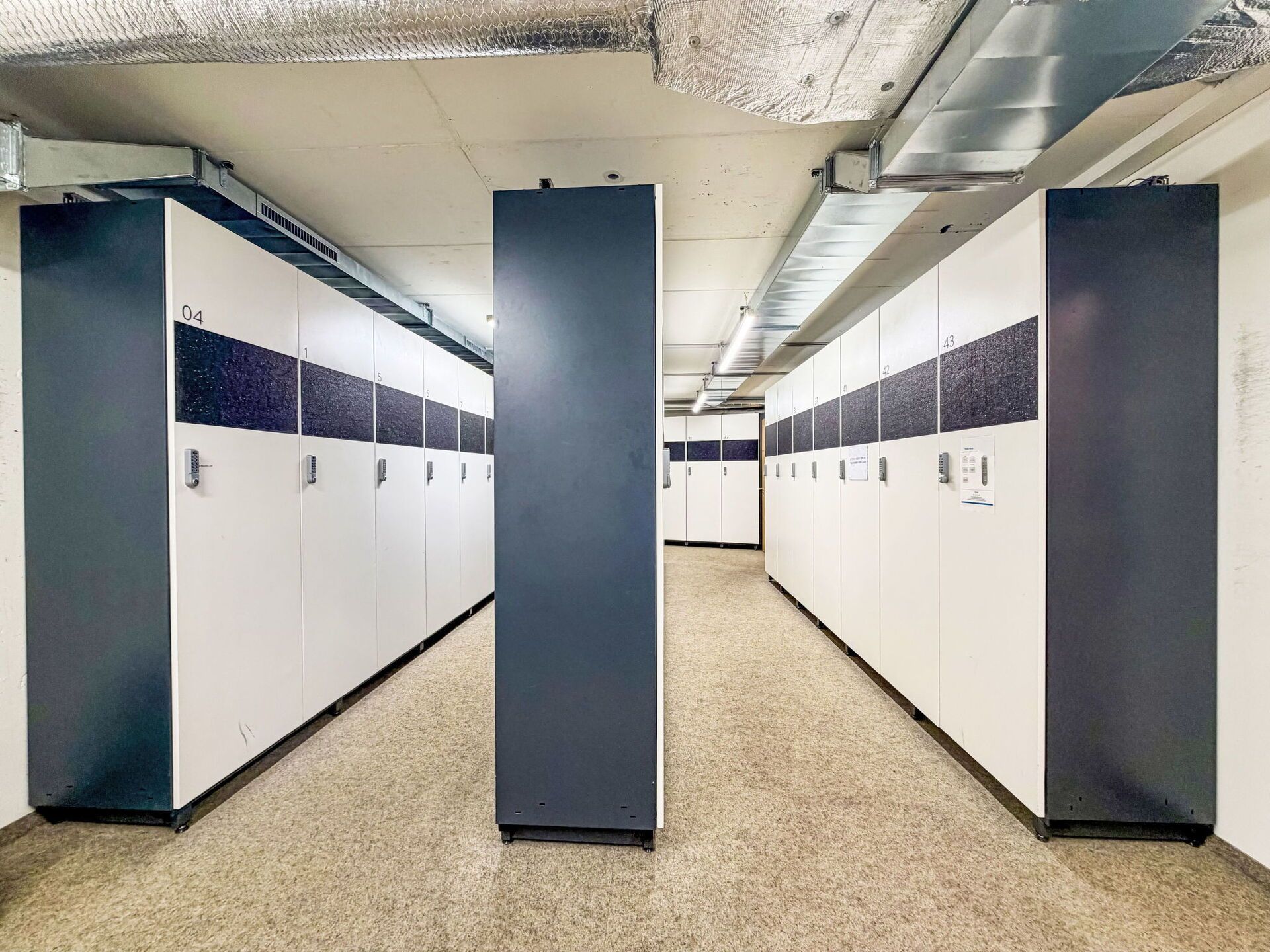 A row of lockers with numbers on the doors, a carpeted floor, and a ceiling with visible ductwork.