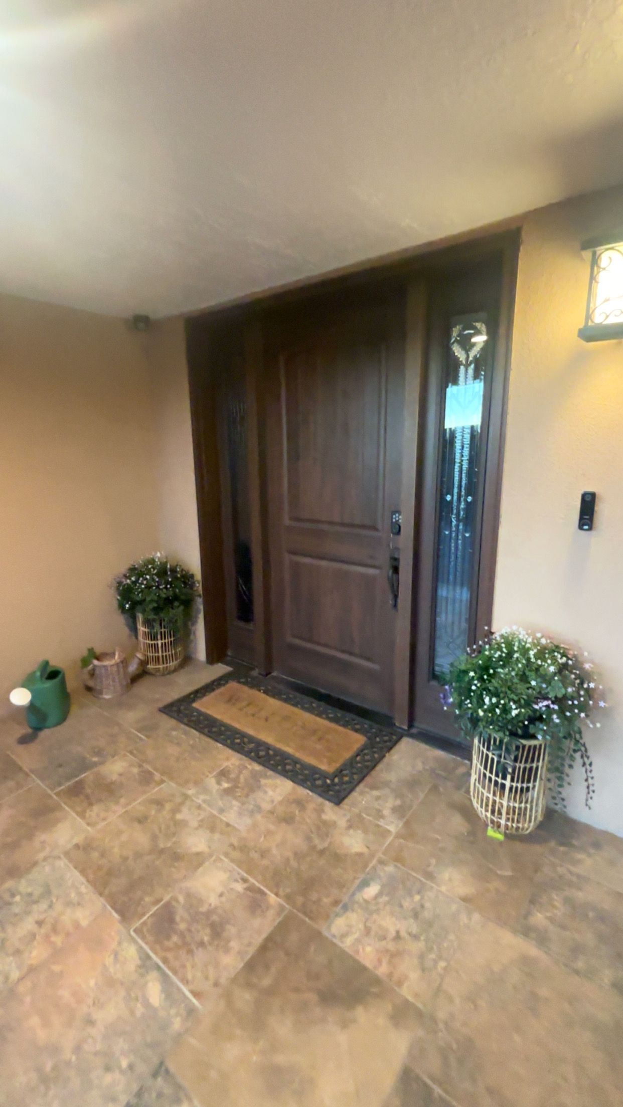 A wooden front door with a doormat, potted plants, and a wall-mounted light fixture.