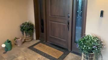 A wooden front door with a doormat, potted plants, and a wall-mounted light fixture.