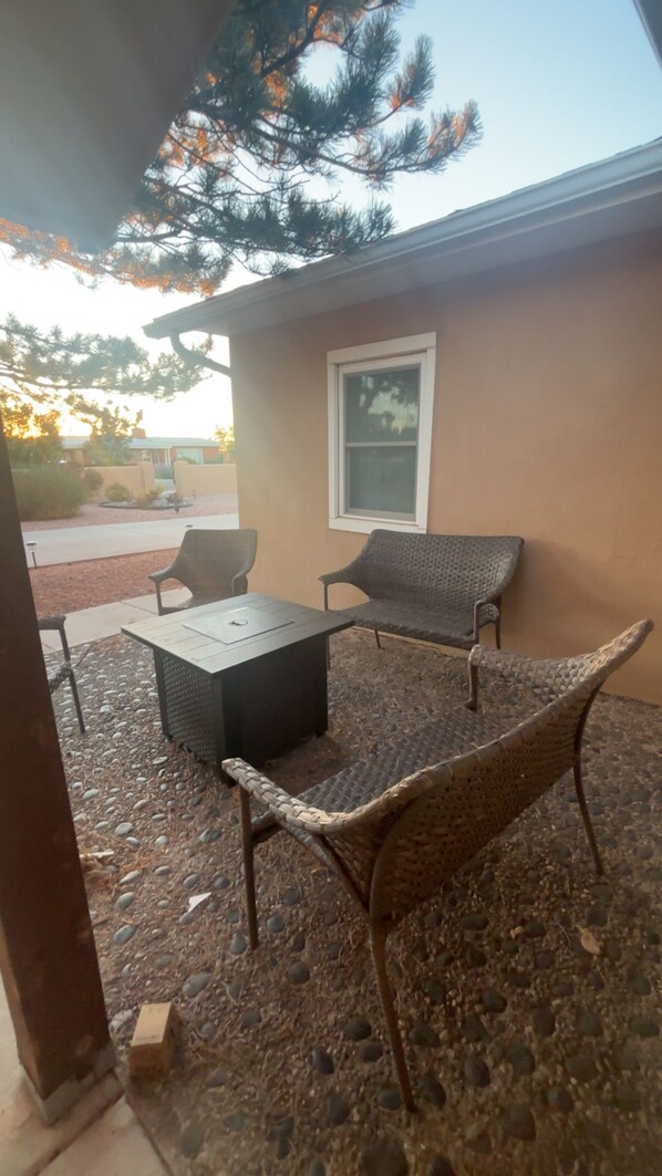 An outdoor patio with a table and two chairs, a window, and a tree in the background.