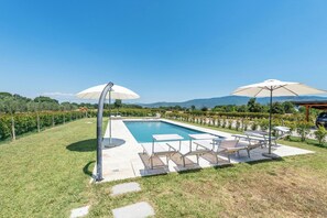 Ein Swimmingpool mit Liegestühlen und Sonnenschirmen, umgeben von einem angelegten Garten und Blick auf ferne Hügel.
