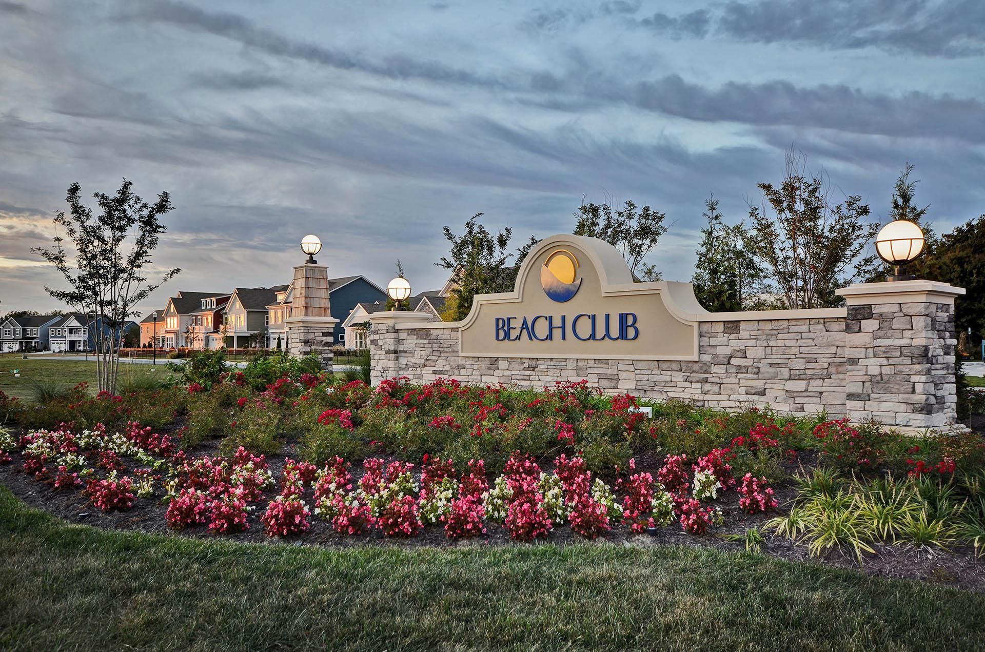 A sign for Beach Club with a stone wall and a landscaped garden with pink flowers.