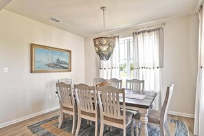 A dining area with a wooden table and chairs, a hanging light fixture, a painting on the wall, and a window with curtains. - Beautiful OVBC townhouse steps from the Amenities! Wifi, Pool, Fitness Center, Sauna, Weekend Shuttle! (Ocean View)