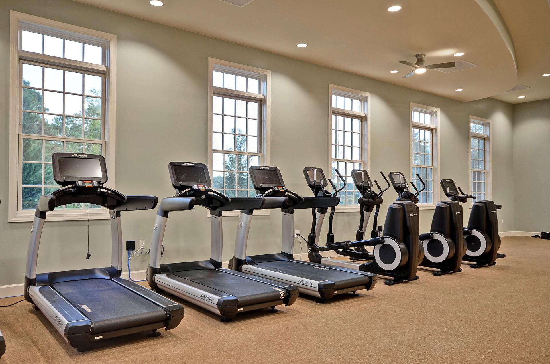 Une salle de remise en forme équipée de tapis de course et d’appareils elliptiques, dotée de grandes fenêtres et d’un ventilateur au plafond.