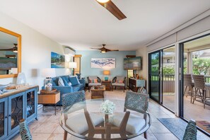 A spacious living room with a glass coffee table, blue sofa, and a dining area with wicker chairs. - Remodeled Ocean View | Wailea Ekolu 703 (Wailea)