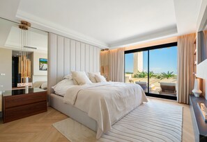 Ein modernes Schlafzimmer mit einem großen Bett, einem Nachttisch, einem Spiegel und Blick auf den Strand.